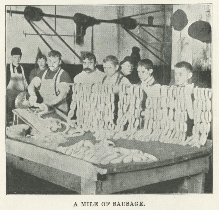 Black-and-white photo of white men and boys processing sausage. A man removes the sausage from a processing machine, and the boys hang it over a long rod.