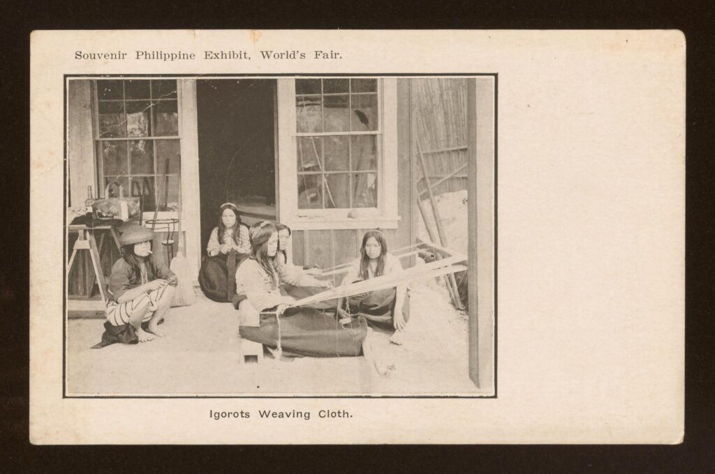 Postcard with black-and-white photo showing five Cordilleran Indigenous women sitting on the ground of a porch. Two weave cloth on backstrap looms and another has a small fiber craft in her hands.