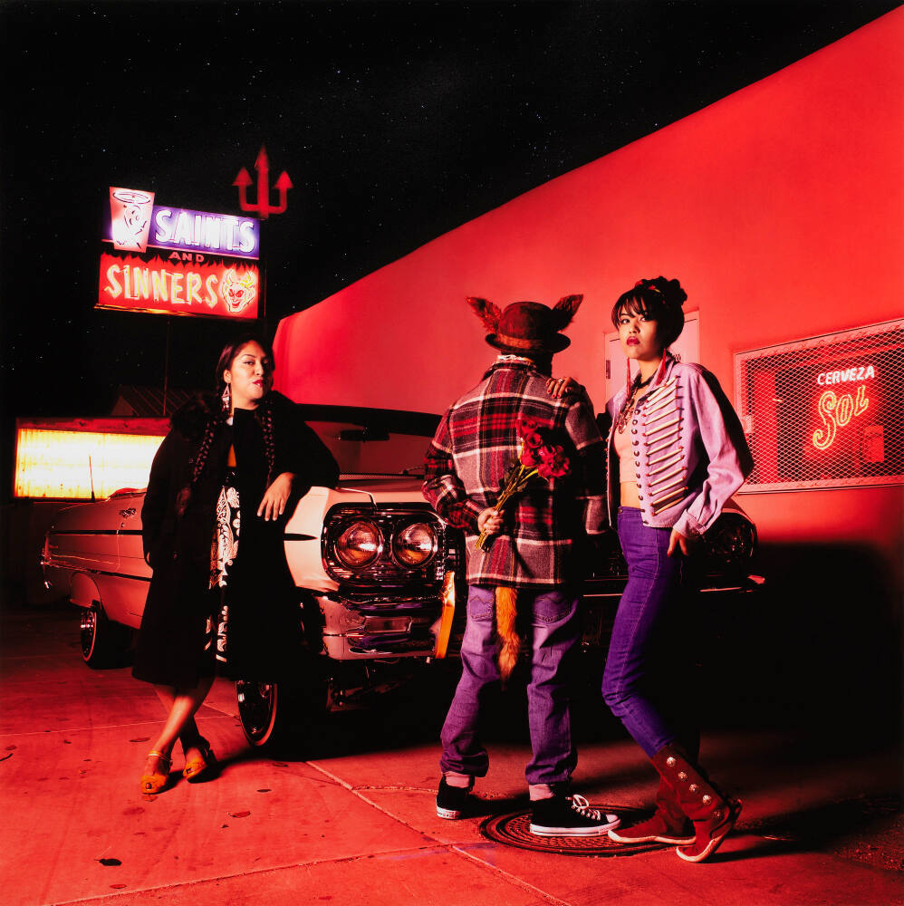 Color photograph of three people standing outside a building at night. The bar has a large neon sign in the upper left reading "Saints/Sinners" with pictures of an angel, a devil, and a pitchfork. A vintage car is parked alongside the building, and an Indigenous woman wearing braids, earrings, and a long black coat leans against it. To her left at the front of the car are two other figures. First, someone with their back to the camera wearing a coat, hat, and jeans. They have coyote ears and a tail and carry a bouquet of roses behind there back. An Indigenous woman stands facing the camera with her hand on the figure's shoulder. She wears her hair up and earrings, a cropped jacket, skinny jeans, and boots. The whole image is tinted with red light.