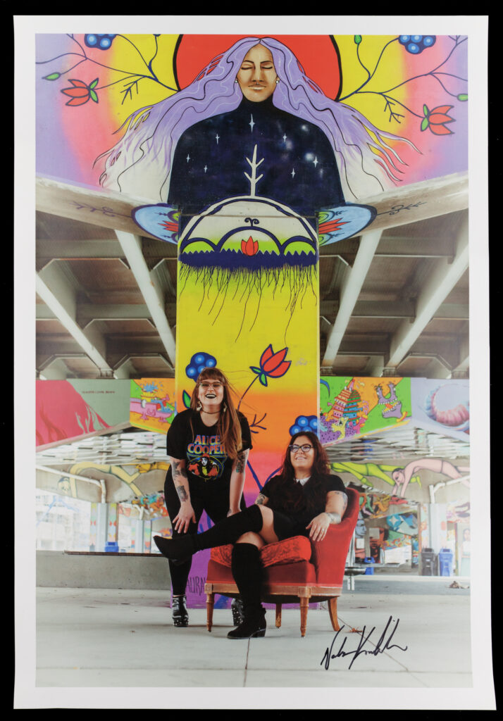 Color photograph of two Indigenous women posing under a concrete overpass decorated with murals. One woman sits in a red armchair and the other stands behind her leaning down with her hands on her knees. Both wear black. The mural behind them shows a brown-skinned woman with purple hair and a serene expression rising out of a landscape.