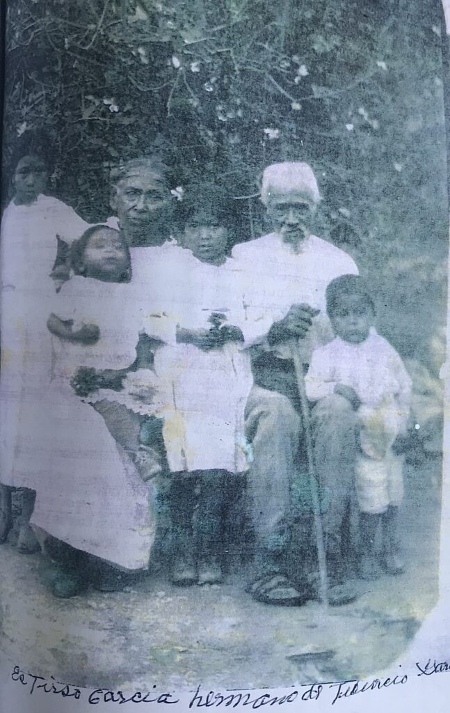 Black-and-white image of a group of Indigenous elders and children. An elderly woman and man sit in front of a wall of vines. Each has a small child on their lap. A slightly older child stands between them, and a fourth child stands to the woman's side.