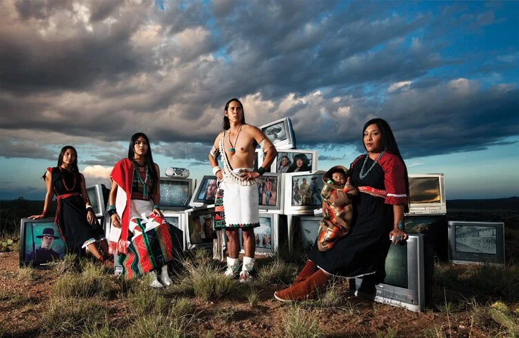 Four Indigenous adults and a baby wearing traditional clothing stand and sit around a pile of vintage TVs playing scenes from old Westerns. They are outdoors in a prairie landscape with dramatic clouds overhead.