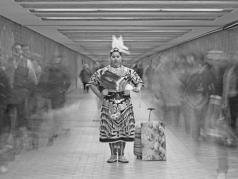 Black-and-white photo of an Indigenous woman in regalia and holding a feather fan in the center of a pedestrian tunnel. She has a suitcase beside her. On either side of her are the blurred forms of other people passing through the tunnel around her.