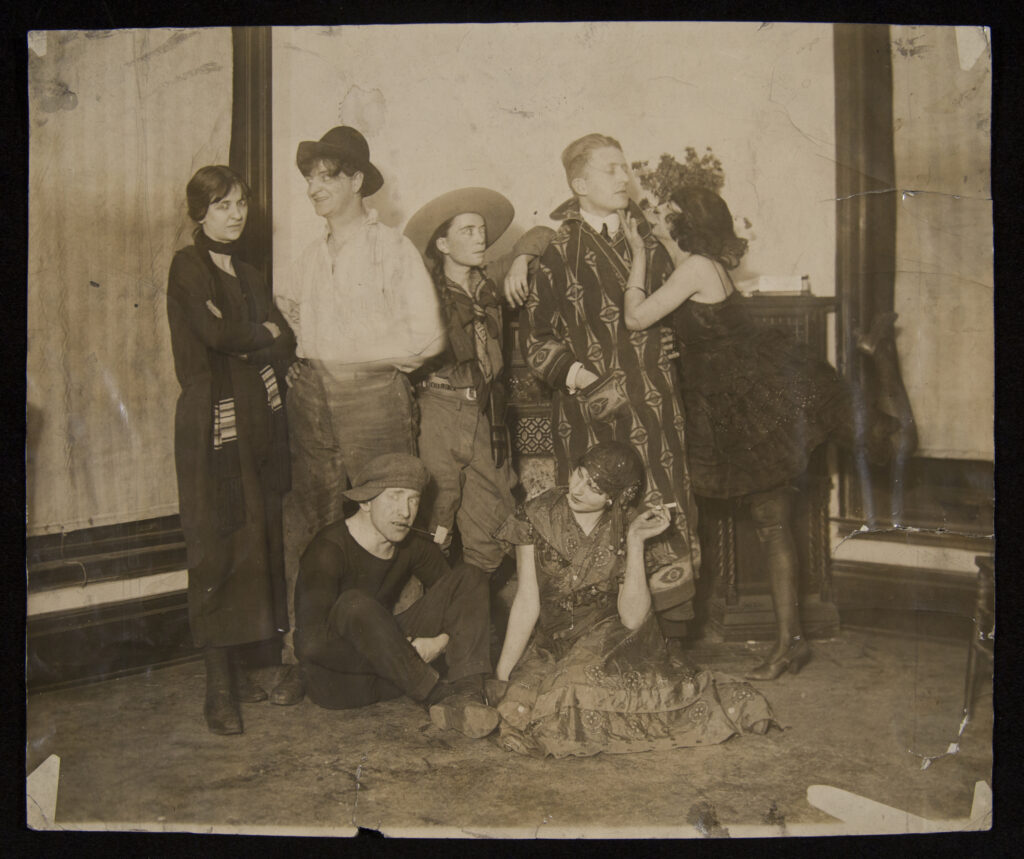 Black-and-white photo of seven people posing in costumes in a shabby room.