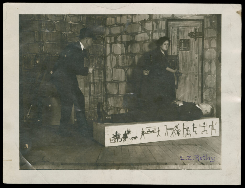 Black-and-white photograph of three people on a low stage. A man holding a gun walks toward a woman opening a door and a man laying on a block decorated with pictographic illustrations.