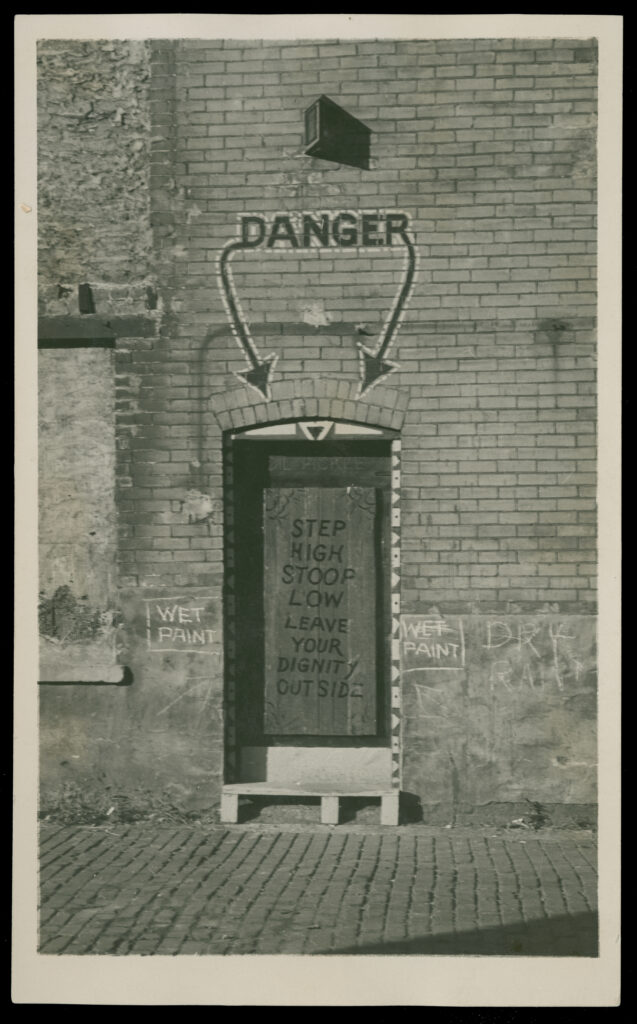 Black-and-white photograph of a door in an alley. The word "Danger" is painted above with arrows pointing down towards the door. On the door is written "Step high stoop low leave your dignity outside."