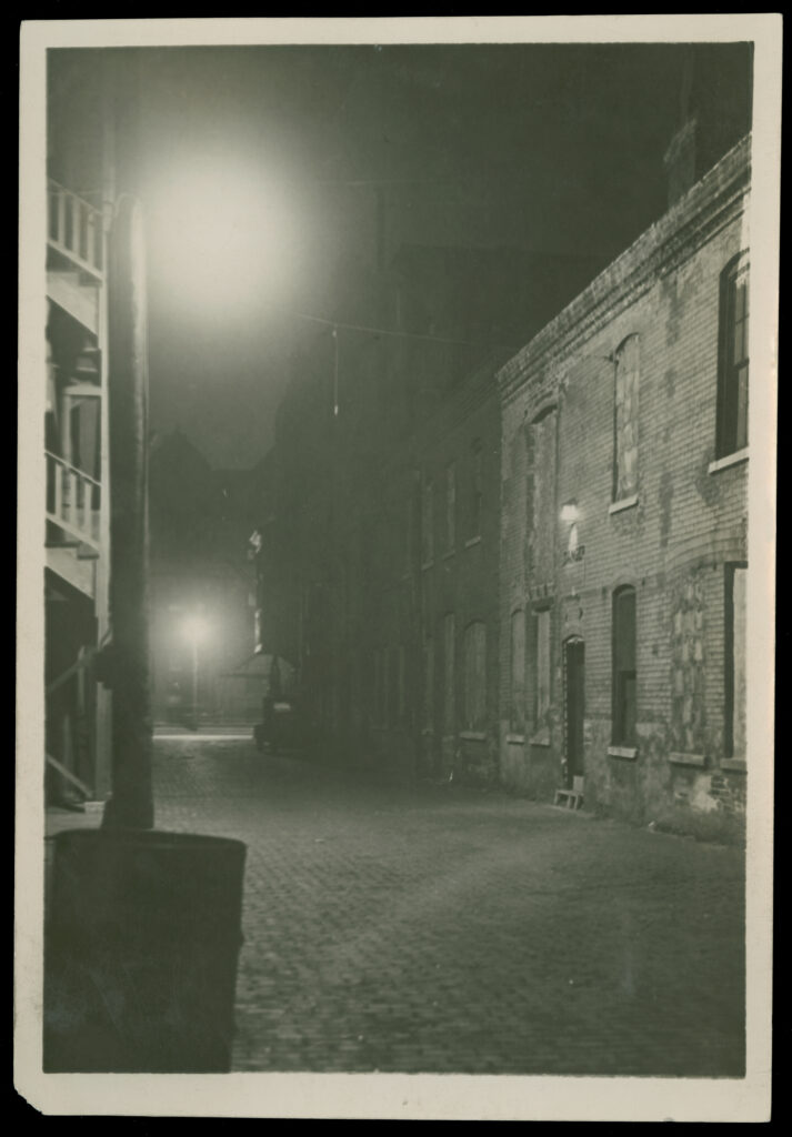 Black-and-white photo of a nighttime view of an alley in a city. On the left are buildings with brick walls, on the right buildings with wooden fire escapes. The scene is illuminated by streetlights.