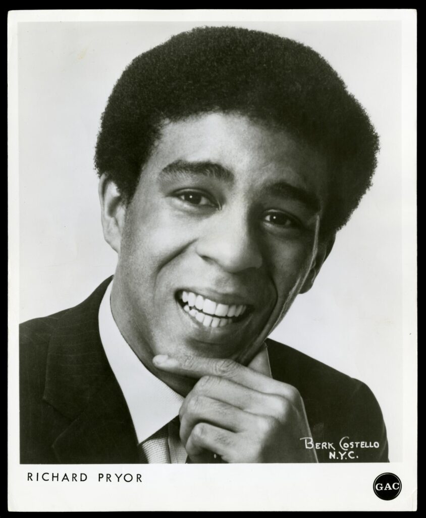 Black-and-white photograph of Richard Pryor, a young Black man wearing a suit. The photograph is taken from the chest up. Pryor is smiling and has his hand on his chin in a thoughtful pose.