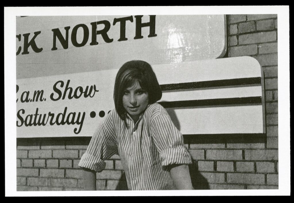Black-and-white photograph of Barbara Streisand, a young white woman with dark shoulder-length hair wearing a striped collared shirt, posing in front of a brick wall. A sign partly visible behind her advertises her show at Mister Kelly's/