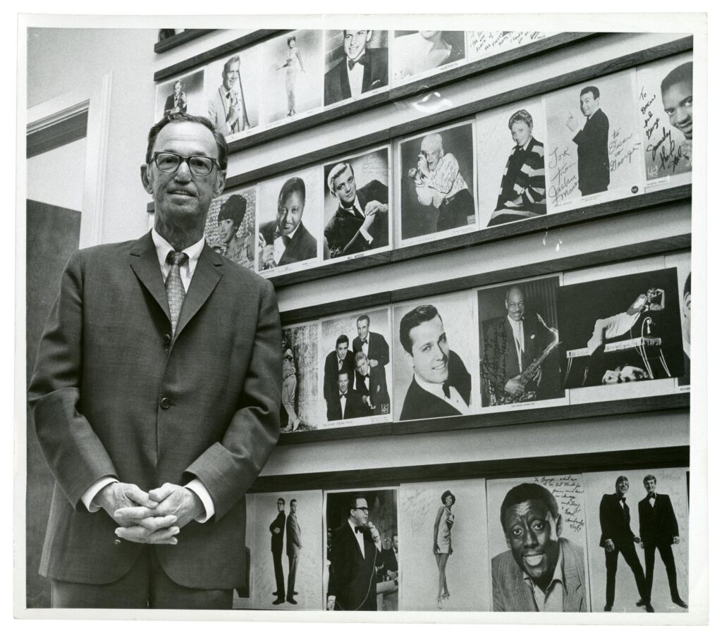 Photograph of George Marienthal, a middle-aged white man wearing a suit, in front of a wall of signed photographs of people who performed at Mister Kelly's.