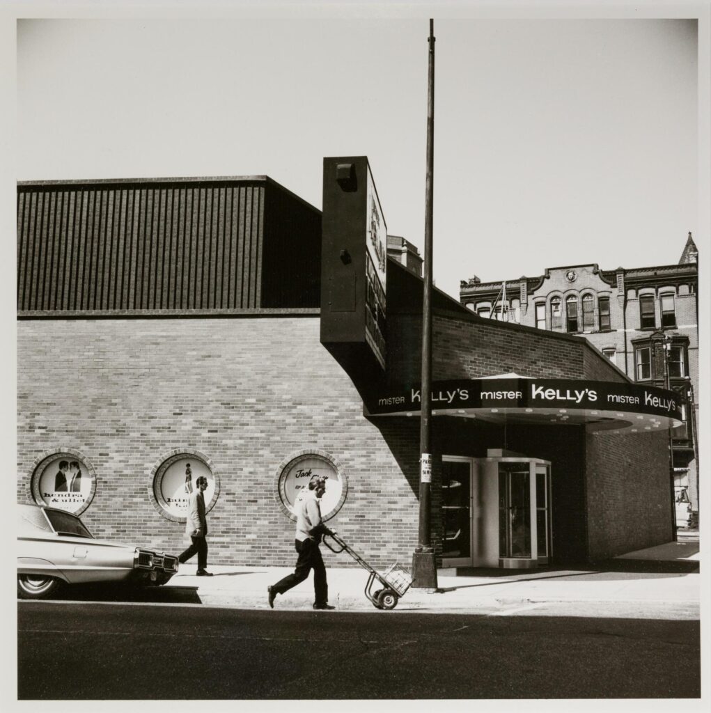 Black-and-white photograph of the exterior of Mister Kelly's, a nightclub with midcentury modern architecture on the corner of a city street. Two men walk by the club, one pushing a small dolly with a crate.