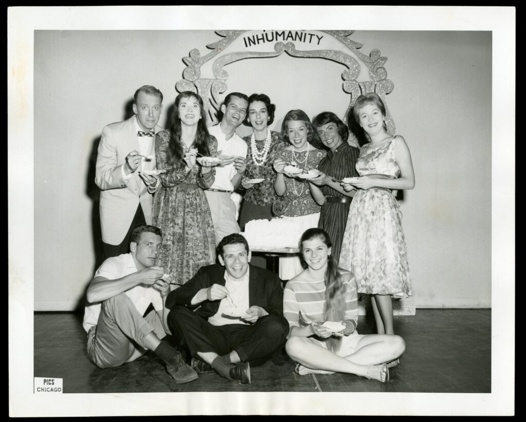 Black-and-white photograph of a group of well-dressed young white people standing and sitting in front of a sparkling sign. Only the bottom of the sign is visible in the photo, and it reads, "inhumanity." There is a cake on a small table in the center of the group, and everyone has a slice.