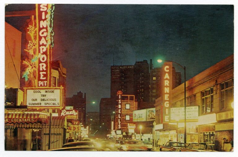 Color postcard showing a city street at night. The street is lined with low buildings lit with bright lights and signs advertising restaurants, bars, and entertainment.