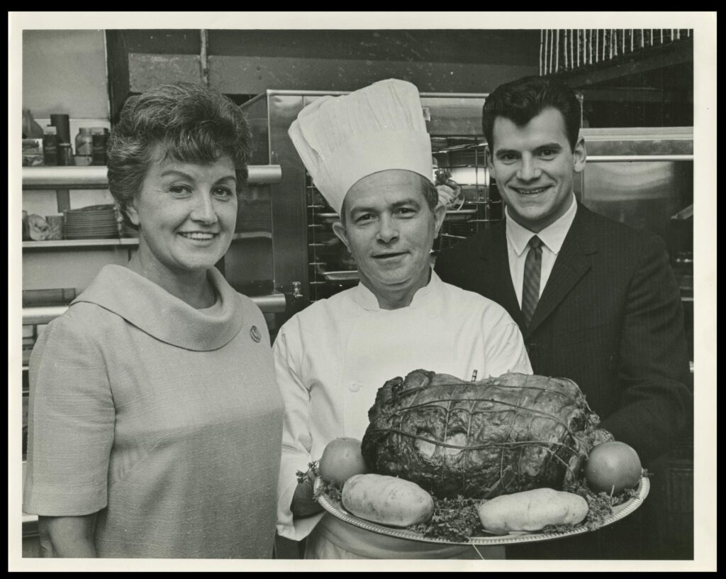 Black-and-white photograph of three white adults in a kitchen, two men and a woman. The woman and the man on the right are dressed in business clothing, while the man between them is dressed as a chef and holds a pot roast on a platter.