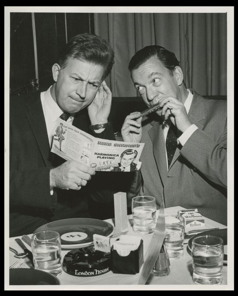 Black-and-white photograph of two white men sitting at a restaurant table. One plays the harmonica while the other looks at a booklet about how to play the harmonica and listens with a concerned look on his face.