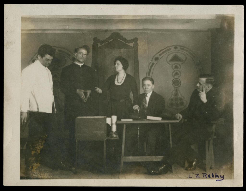 Black-and-white photograph of five people in a row on stage. Two men stand at stage right. In the center stands a woman. Two men sit at stage left. Behind them is a painted backdrop.